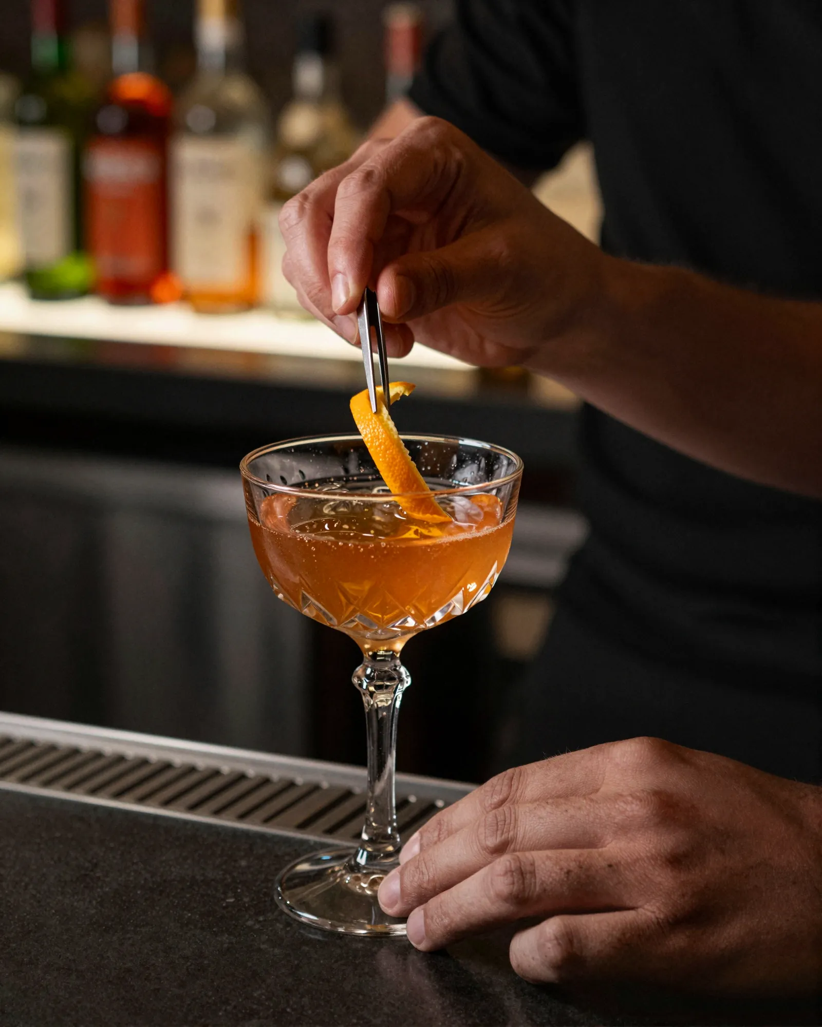 A bartender’s hands placing an orange peel twist with tweezers on an amber cocktail