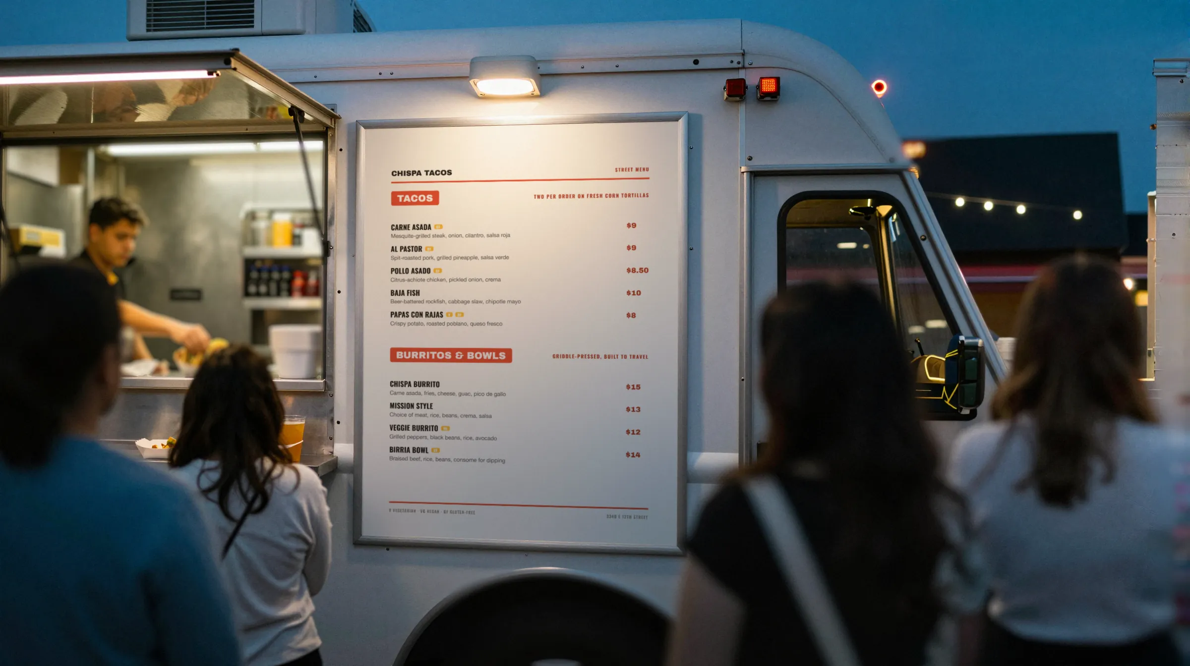 A food truck service window at dusk with a printed menu board beside it and a line of customers