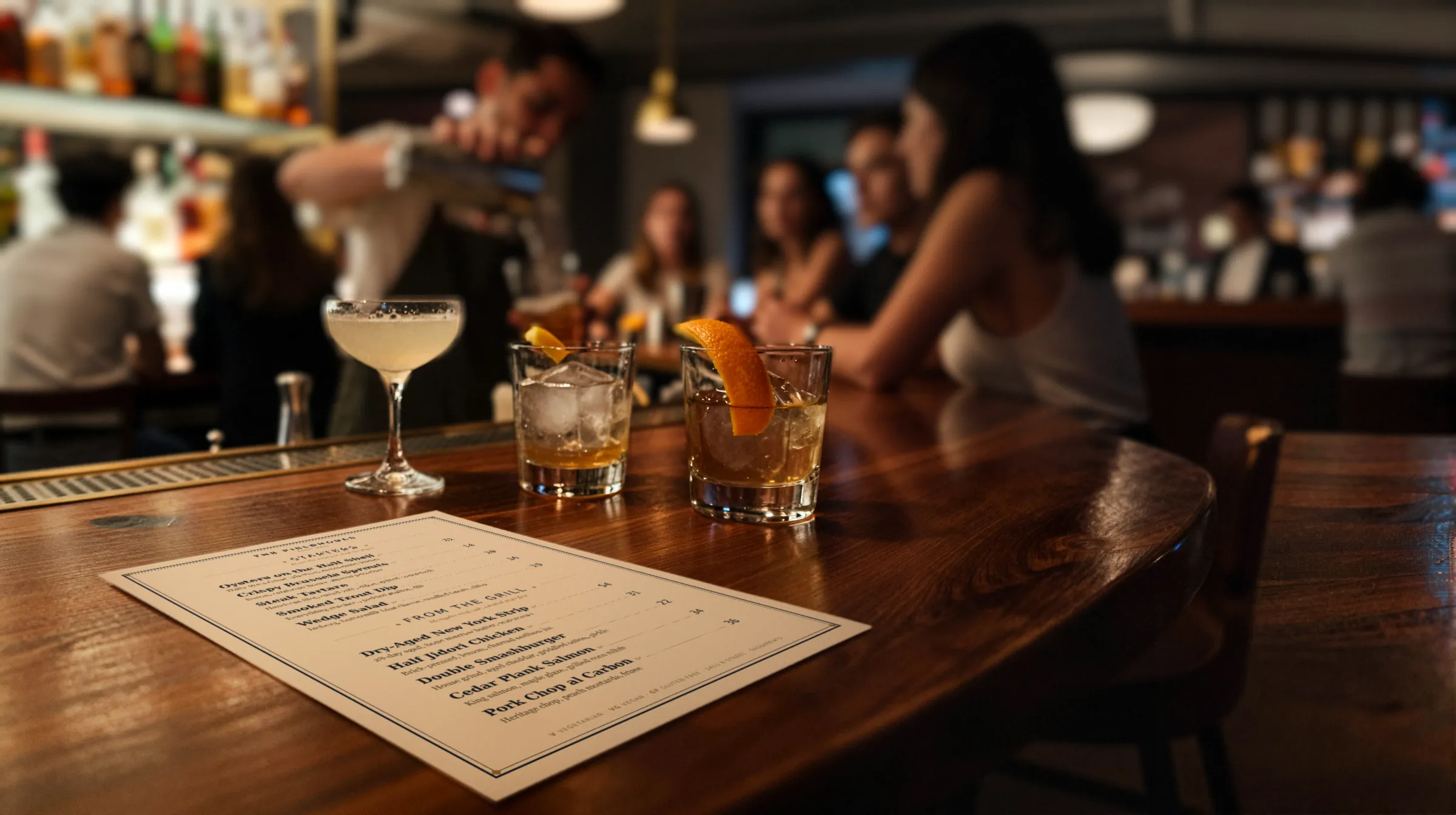 A busy cocktail bar at night, a printed menu on the counter beside a coupe and two old fashioneds