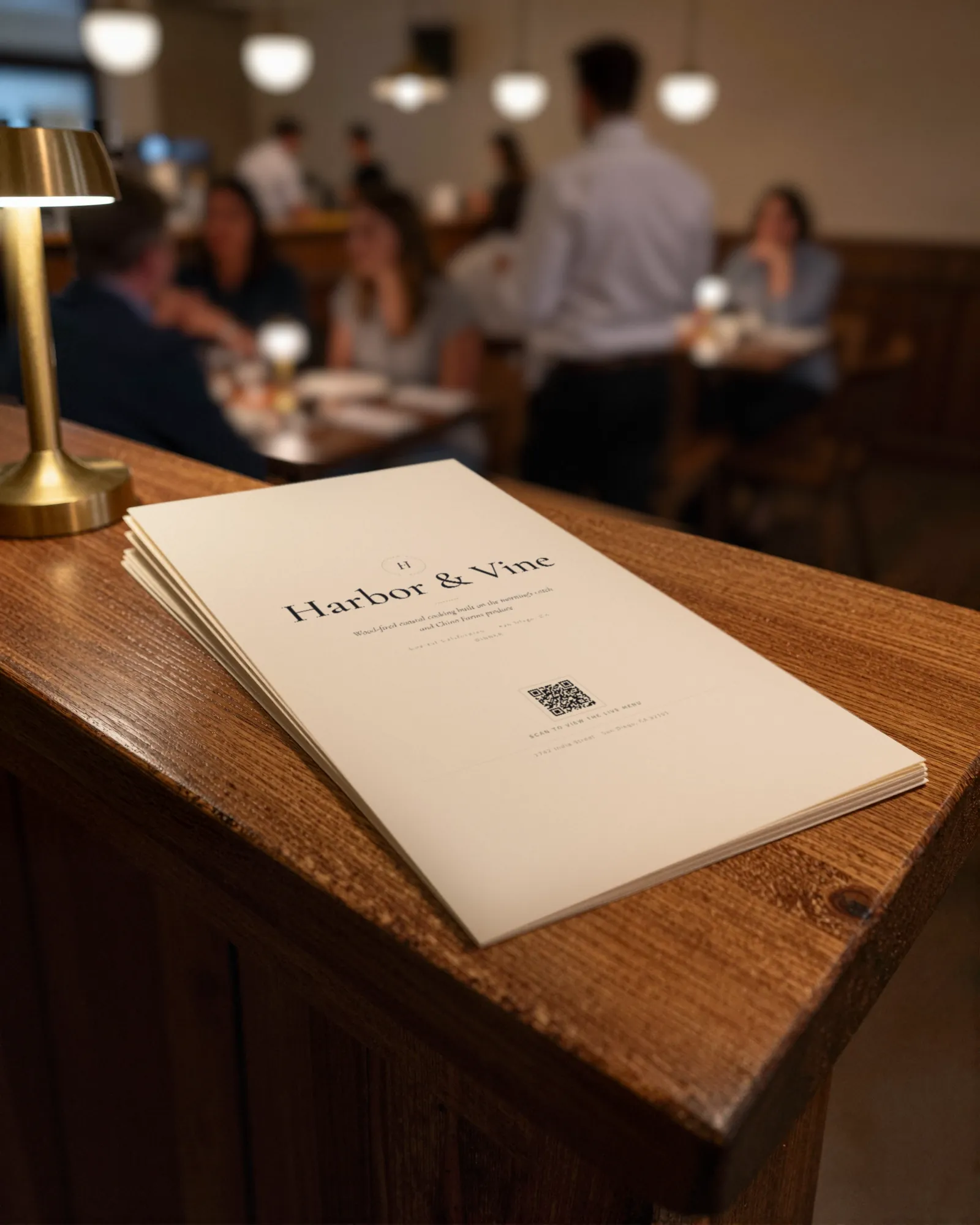 A stack of printed menus on a wooden host stand beside a brass lamp, a busy dining room blurred behind